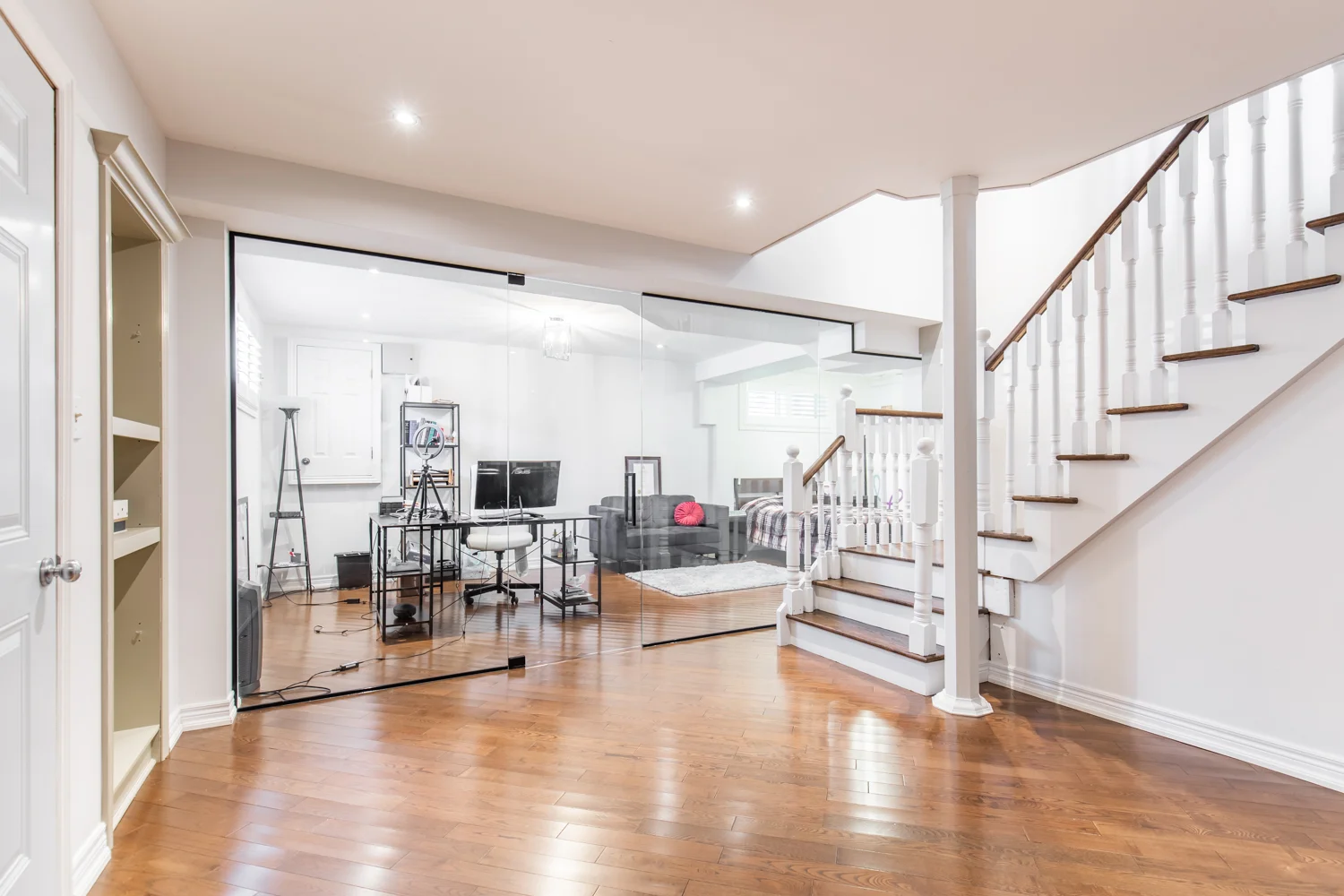 photo-of-basement-room-with-hardwood-floor-glass-partition-office-desk-equipment-and-white-staircase-leading-upstairs