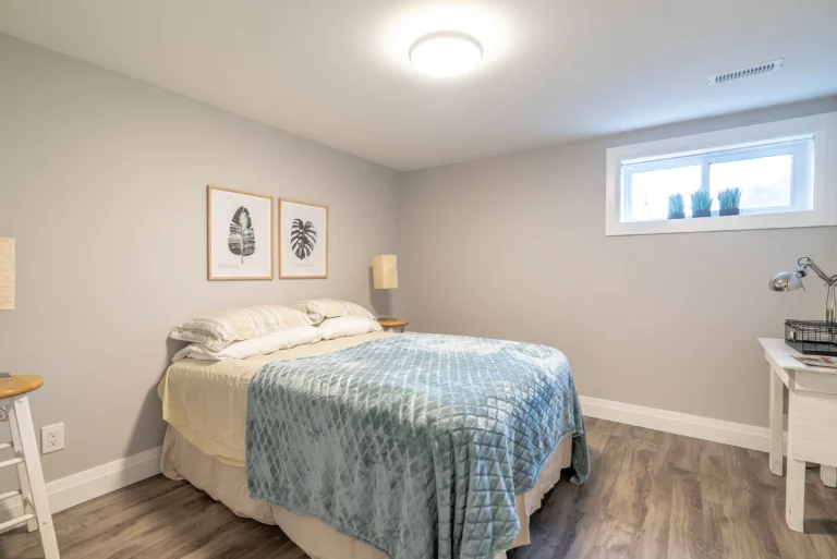 photo-of-bedroom-with-bed-blue-blanket-framed-leaf-art-wall-lamp-small-window-and-wood-floor