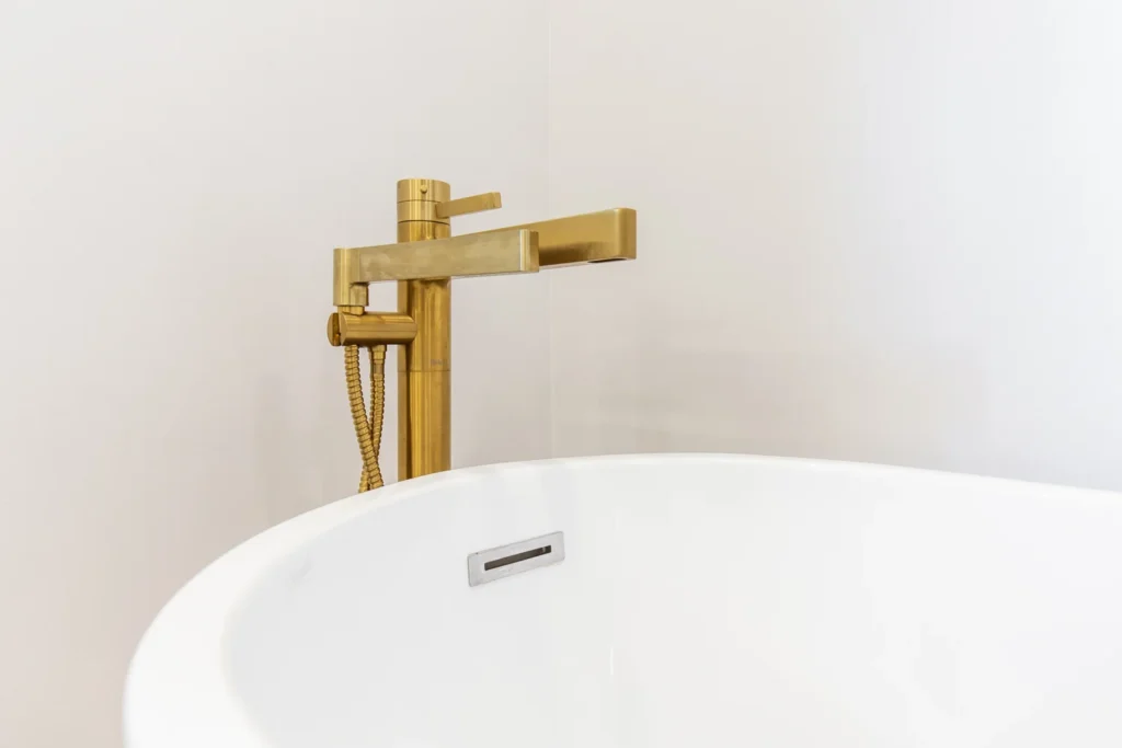 photo-of-gold-freestanding-bathtub-faucet-with-hand-shower-behind-white-bathtub-against-light-colored-wall