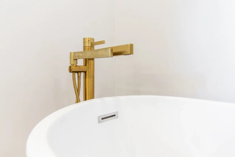 photo-of-gold-freestanding-bathtub-faucet-with-hand-shower-behind-white-bathtub-against-light-colored-wall