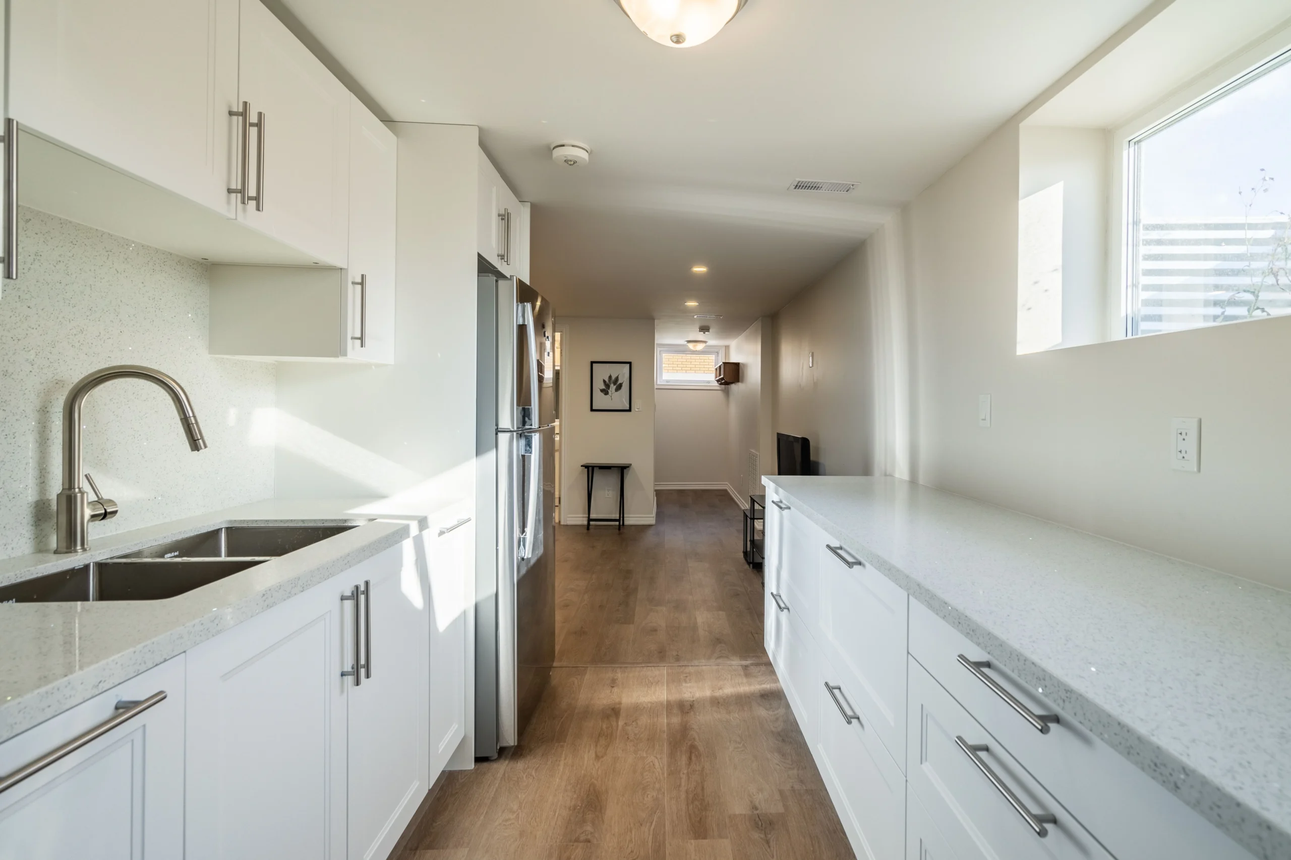 photo-of-long-white-kitchen-with-stainless-steel-refrigerator-double-sink-cabinets-and-wood-flooring