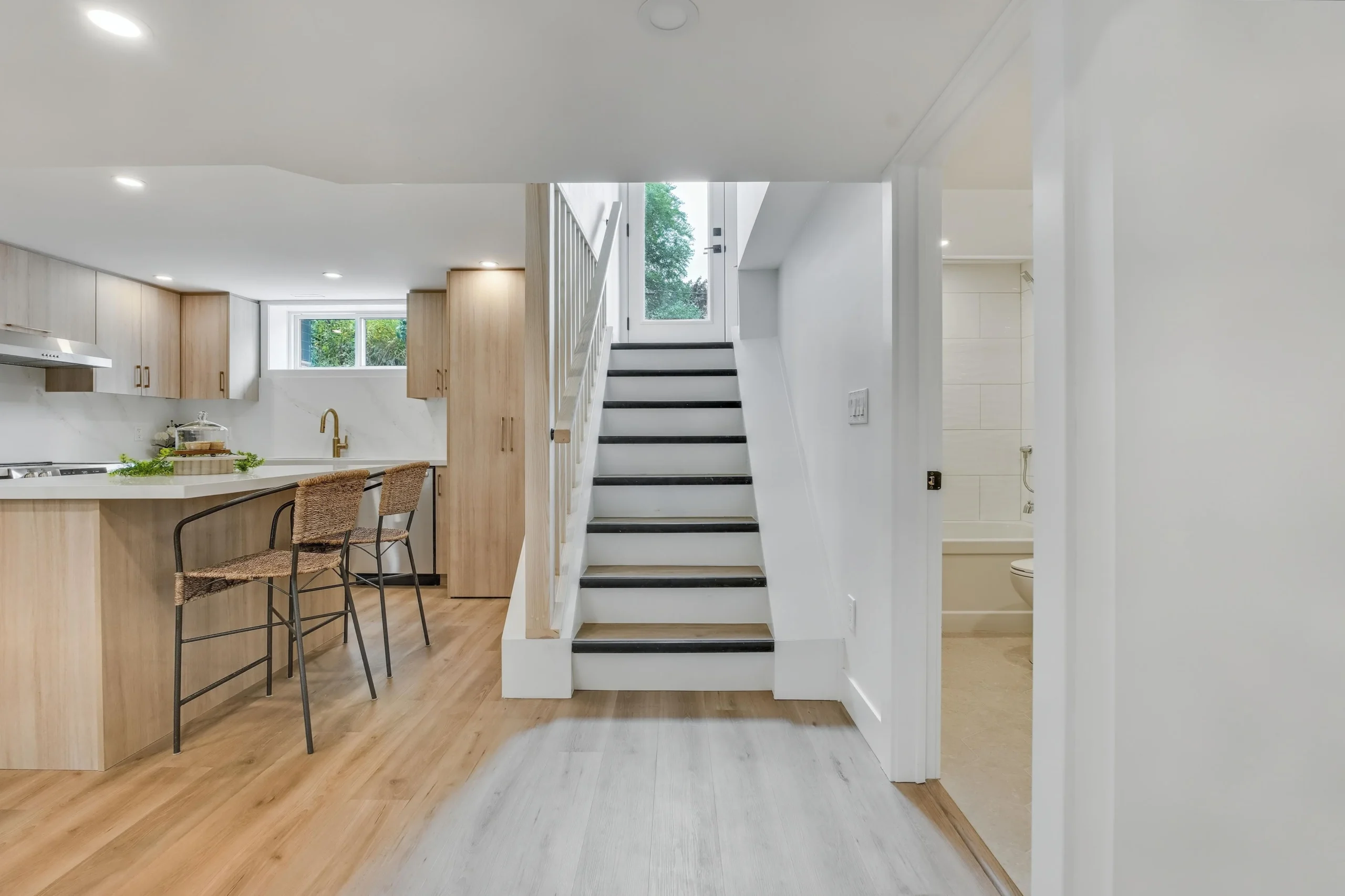 photo-of-modern-basement-kitchen-with-bar-stools-staircase-and-bathroom-showing-home-renovations-ancaster-with-light-wood-cabinets-white-walls-and-recessed-lighting