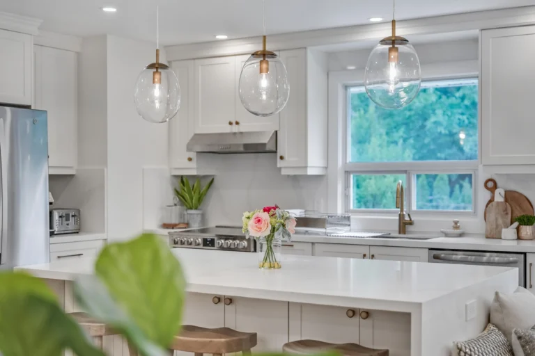 photo-of-modern-white-kitchen-with-island-glass-pendant-lights-window-sink-and-flower-vase-on-counter