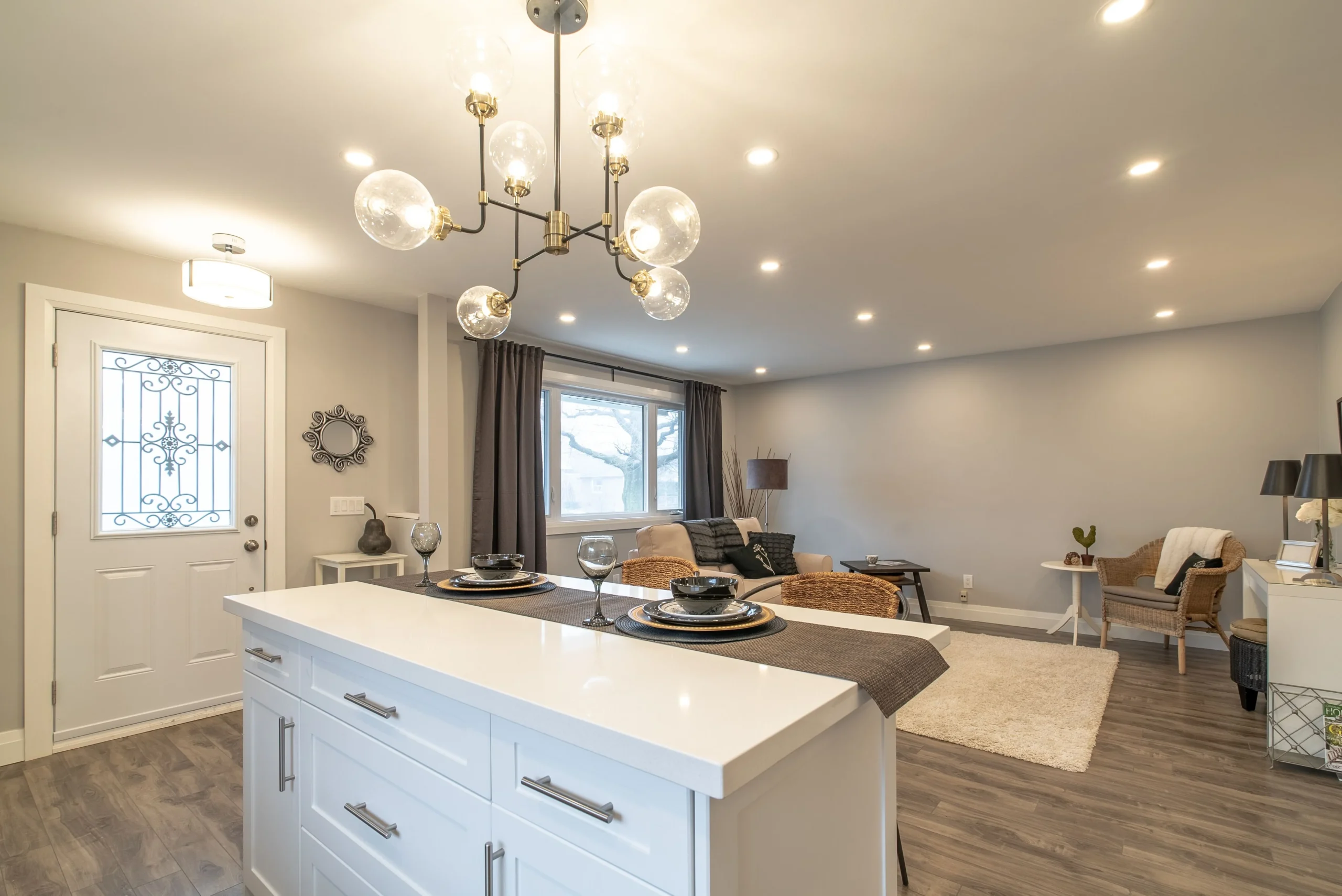 photo-of-open-concept-kitchen-and-living-room-with-white-island-chandelier-front-door-sofa-window-and-recessed-ceiling-lights