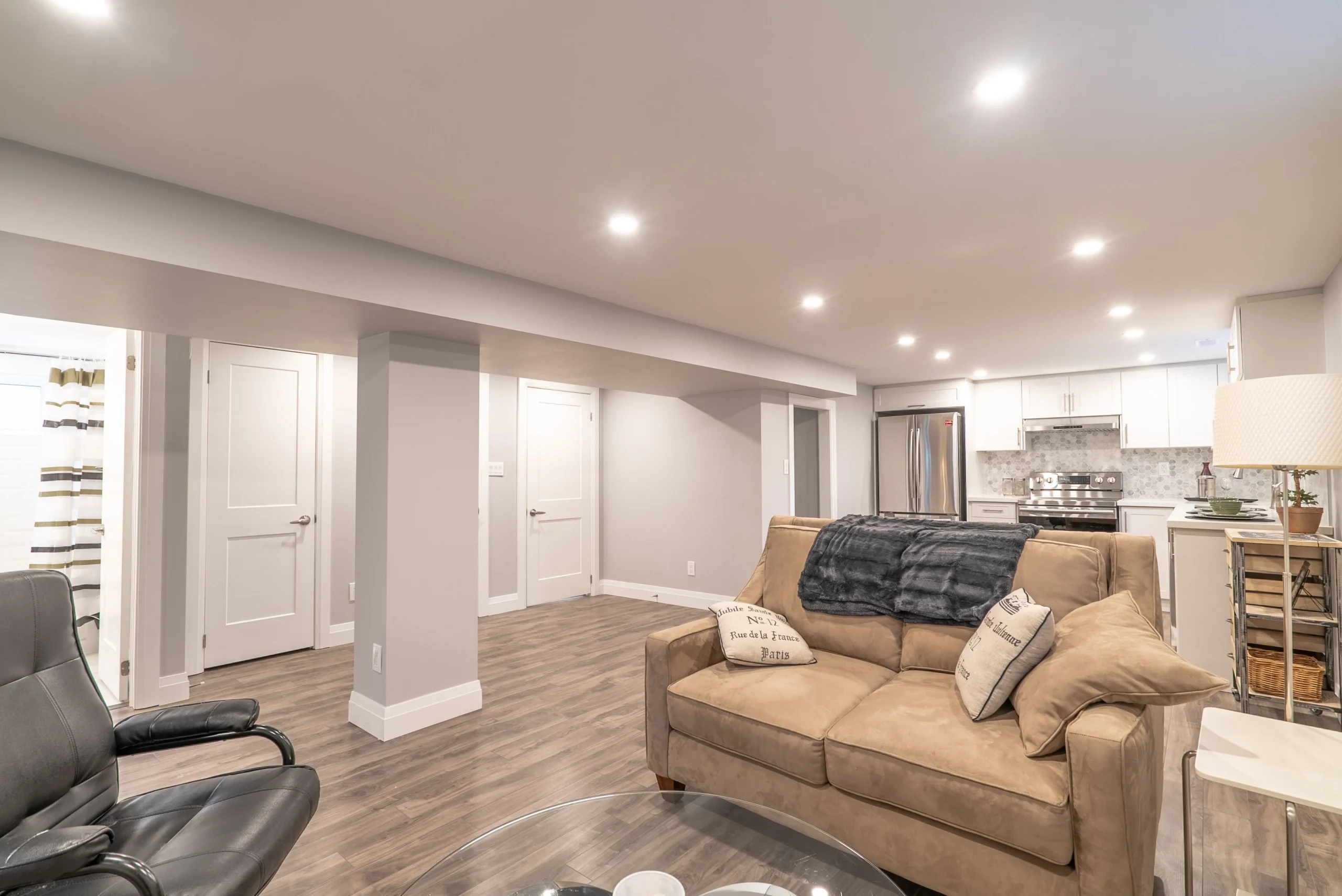 photo finished basement living area with brown sofa black chair coffee table recessed lighting support columns and open kitchen in background