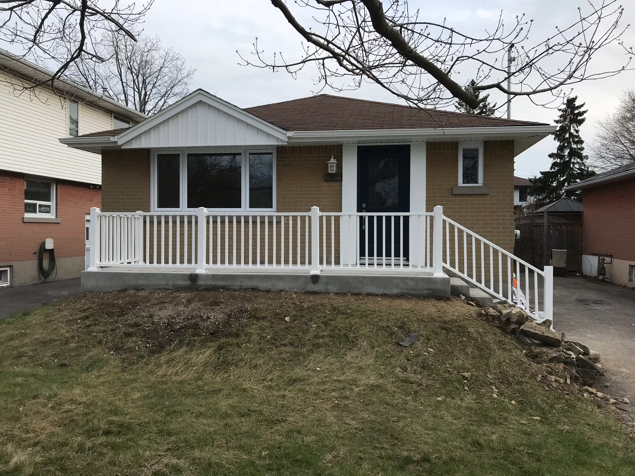 photo single story brick house with white railing front porch black door overcast sky bare trees and sloped yard in suburban neighborhood
