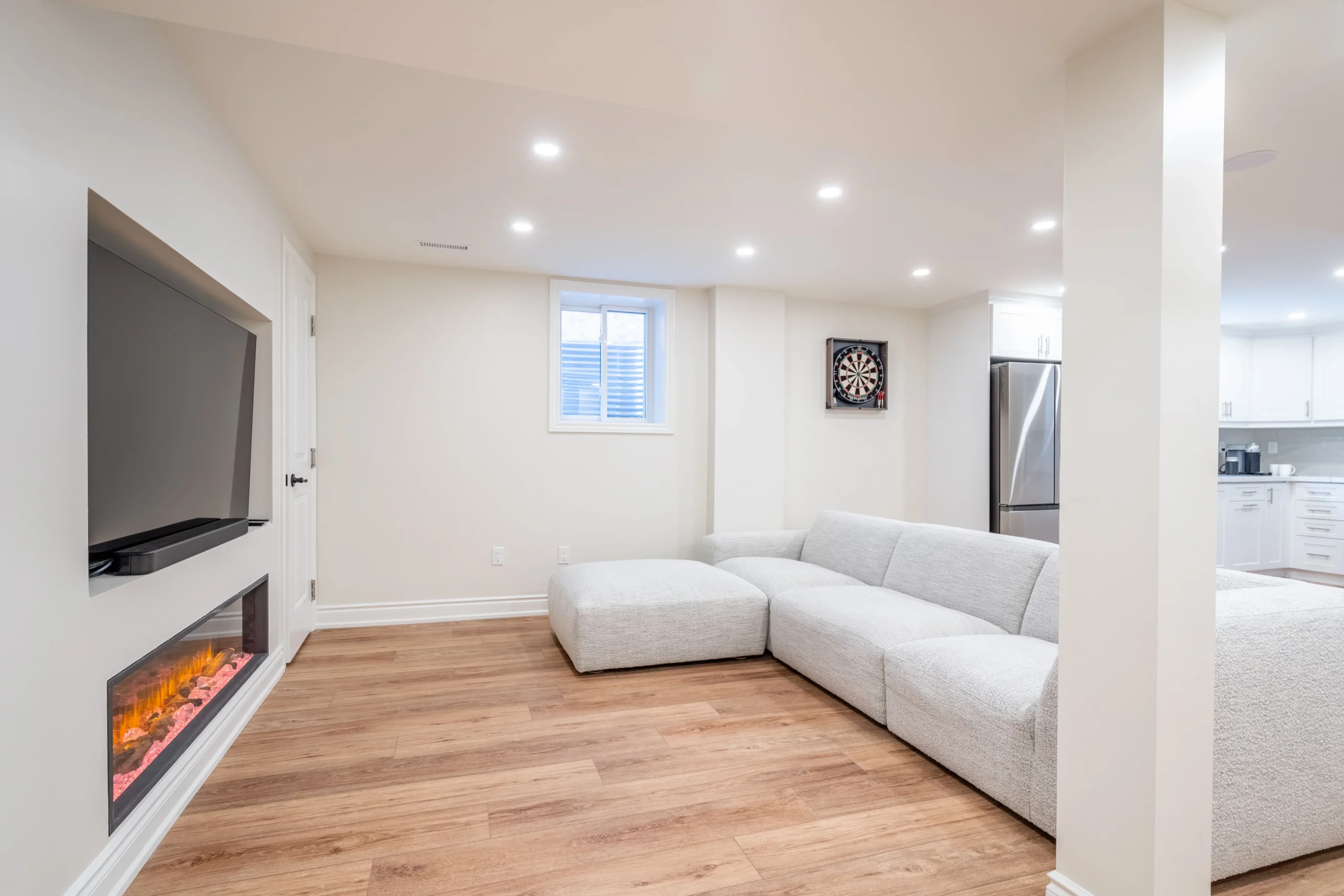 Photo of modern basement living room with sectional sofa, fireplace, dartboard, and kitchenette in background