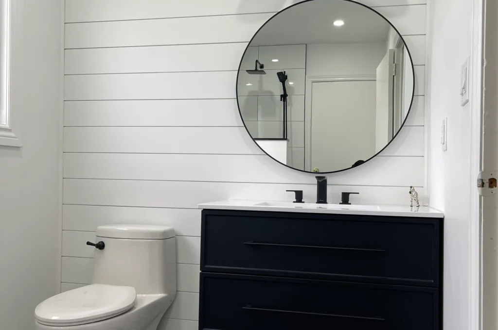 photo modern bathroom with toilet black vanity round mirror three light fixture white shiplap wall black floor tiles and glass shower visible