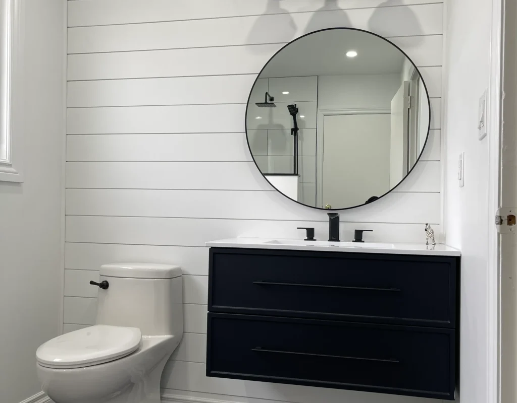 photo modern bathroom with toilet black vanity round mirror three light fixture white shiplap wall black floor tiles and glass shower visible